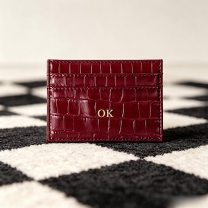 Peut inclure: Un porte-cartes rouge foncé avec une texture en cuir de crocodile brillant. Cet accessoire rectangulaire présente des fentes horizontales pour les cartes et porte les lettres dorées OK gravées au centre de la surface en similicuir.