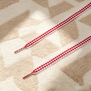 May include: Two flat shoelaces featuring a red and white gingham check pattern. Each lace has a scalloped white edge and a solid red plastic aglet at the tip. The laces are long, narrow, and designed for threading through eyelets on footwear.