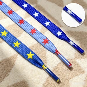 May include: Three flat shoelaces with star patterns. One is dark blue with white stars, one is light blue with red stars, and one is dark blue with yellow stars. Each lace features a clear plastic aglet at the end, with a circular inset showing detail.