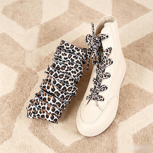 Könnte beinhalten: Ein hoher Sneaker in einfarbigem Creme mit abgerundeter Zehenpartie und Gummisohle. Der Schuh verfügt über flache, breite Schnürsenkel mit einem schwarz-braun-weißen Leopardenmuster, die zudem separat aufgerollt neben dem Schuh abgebildet sind.