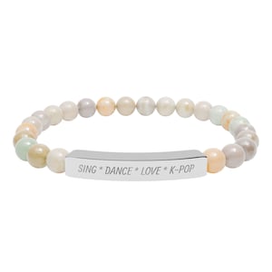 Könnte beinhalten: Ein elastisches Armband mit glatten, pastellfarbenen Perlen in Creme, Pfirsich, Hellblau und Grau. Ein zentraler, polierter silberfarbener Metallsteg ist mit dem Text SING * DANCE * LOVE * K-POP graviert.
