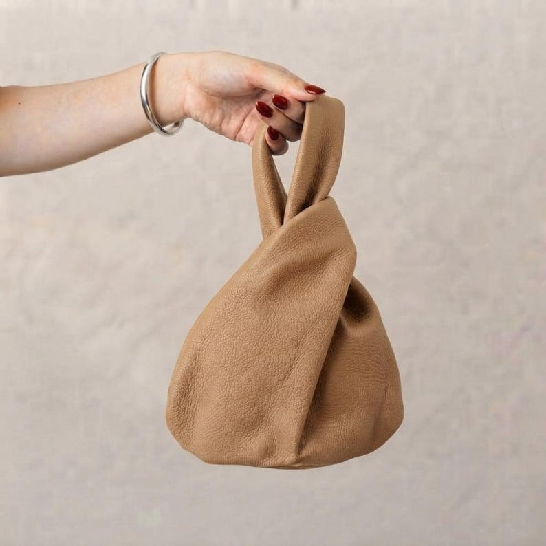 K&ouml;nnte beinhalten: Eine beigefarbene Handtasche aus strukturiertem Leder mit einem knotenartigen Griffdesign, bei dem eine Seite durch die andere gezogen wird. Die Tasche hat eine weiche, lockere Silhouette und einen gleichm&auml;&szlig;igen hellbraunen Farbton auf der Oberfl&auml;che.