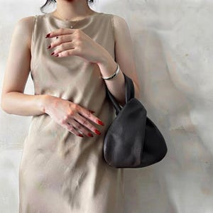 K&ouml;nnte beinhalten: Eine dunkel anthrazitfarbene Lederhandtasche mit geknotetem Griffdesign und einer weichen, genarbten Textur. Die Tasche hat eine abgerundete, lockere Silhouette und wird an einem Griff gehalten, der oben einen Knoten bildet, was ein schlichtes Design unterstreicht.