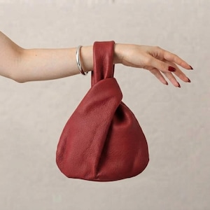 K&ouml;nnte beinhalten: Eine dunkelrote Handtasche aus Leder mit strukturierter Oberfl&auml;che und einem knotenartigen Griffdesign. Die Tasche hat eine weiche, lockere Struktur mit einer einzelnen integrierten Griffschlaufe, die f&uuml;r eine geraffte Optik oben sorgt.