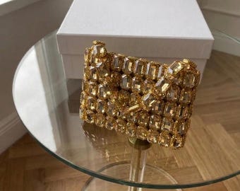 Könnte beinhalten: Eine rechteckige Clutch, die mit facettierten, hellgoldfarbenen Kristallen in einem Gittermuster bedeckt ist. Die Tasche verfügt über einen strukturierten Klappenverschluss und einen metallischen goldfarbenen Verschluss vor einer schlichten weißen Schachtel.