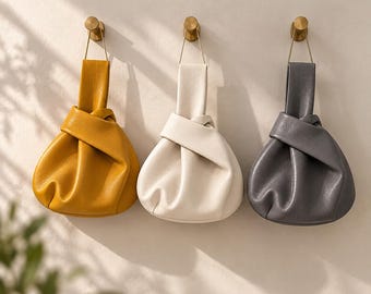 Könnte beinhalten: Drei knotenförmige Handtaschen in Senfgelb, Off-White und Anthrazitgrau. Jede Tasche verfügt über einen integrierten Griff, eine weiche, geraffte Silhouette und eine strukturierte Oberfläche, aufgehängt an einzelnen goldfarbenen Wandhaken.