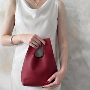 K&ouml;nnte beinhalten: Eine dunkelrote Handtasche mit einer strukturierten, genarbten Oberfl&auml;che und einem ausgeschnittenen Griffdesign. Die Tasche hat eine strukturierte, konische Silhouette mit einer weiten &Ouml;ffnung oben und besteht aus einfarbigem Material mit sichtbarer Maserung.