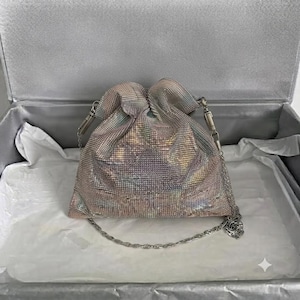 Könnte beinhalten: Eine beutelförmige Handtasche, die mit irisierenden, lichtreflektierenden Kristallen in Rosa-, Blau- und Silbertönen besetzt ist. Die Tasche verfügt über einen gerafften Verschluss an der Oberseite und einen dünnen, abnehmbaren Schulterriemen aus silberfarbener Metallkette.