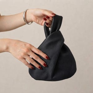 K&ouml;nnte beinhalten: Eine schwarze Handtasche aus Leder mit weicher, genarbter Textur und einem geknoteten Griffdesign. Die Tasche hat eine abgerundete, lockere Form, die sich oben zusammenzieht und ein kompaktes Design zum Tragen kleiner pers&ouml;nlicher Gegenst&auml;nde bietet.