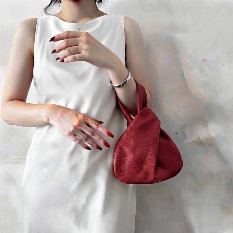 K&ouml;nnte beinhalten: Ein &auml;rmelloses wei&szlig;es Kleid mit hohem Ausschnitt, kombiniert mit einer kleinen Handtasche aus dunkelrotem, strukturiertem Leder. Die Handtasche hat einen geknoteten Griff und eine weiche, lockere Form. Abgerundet wird das Set durch ein schlichtes Silberarmband.