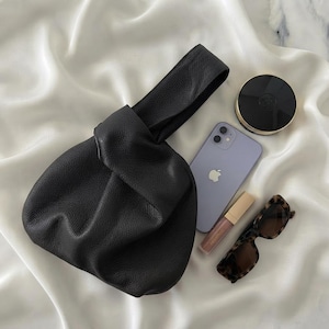 K&ouml;nnte beinhalten: Eine schwarze Handtasche aus genarbtem Leder mit Knotenhenkel, begleitet von einem hellvioletten Smartphone, einem runden schwarzen Puder-Kompakt, einem Lipgloss-Stift und einer braunen Schildpatt-Sonnenbrille auf wei&szlig;em Stoff.