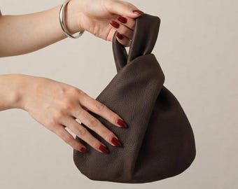 Könnte beinhalten: Eine dunkelbraune Handtasche aus Leder mit strukturierter Oberfläche und einem geknoteten Griffdesign. Die Tasche hat eine weiche, lockere Konstruktion mit einem integrierten Schlaufengriff, der eine geraffte, asymmetrische Form bildet.