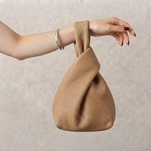 May include: A tan-colored handbag crafted from textured leather, featuring a distinctive knot-style handle design. The bag has a soft, slouchy structure with a single integrated handle loop that creates a gathered appearance at the top opening.