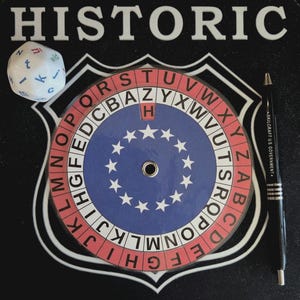 May include: A circular cipher wheel with red and white alphabet rings and a blue centre featuring a circle of white stars. The device is placed on a black mat labelled HISTORIC, accompanied by a white twenty-sided die and a black ballpoint pen.