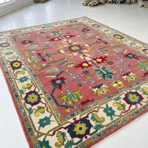 May include: A rectangular area rug featuring a vibrant pink field with a central floral medallion and intricate botanical motifs in shades of teal, yellow, and red. The rug is framed by a wide cream-coloured border with repeating floral patterns.
