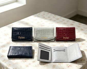 Puede incluir: Una colección de carteras plegables con textura de cocodrilo. Tres carteras están cerradas y muestran el nombre Dylan en letras doradas, mientras que una cartera azul marino está cerrada y una de color gris claro está abierta.