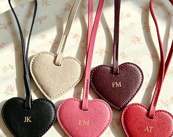 Puede incluir: Cinco etiquetas de equipaje de cuero en forma de corazón con correas largas, cada una con iniciales grabadas en oro. Los colores incluyen negro, beige, burdeos oscuro, rosa brillante y rojo. Cada etiqueta tiene una superficie texturizada.