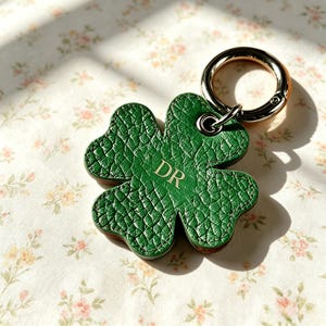 May include: A four-leaf clover shaped keychain made of green textured material with the letters DR embossed in gold. The clover is attached to a polished silver-toned metal ring with a spring-loaded clasp mechanism for secure attachment.