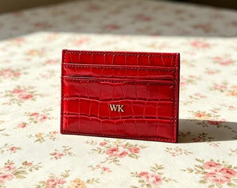 Puede incluir: Un tarjetero rectangular de color rojo con una textura brillante de imitación de cocodrilo. La parte frontal presenta varias ranuras horizontales para tarjetas y las letras doradas WK grabadas en el centro de la superficie inferior.
