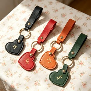 May include: Four heart-shaped keychains with leather straps in black, red, orange, and dark green. Each keychain features a gold-tone metal ring, a gold-tone rivet, and gold embossed initials: JK on black, BH on red, YT on orange, and MV on green.