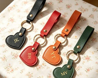 May include: Four heart-shaped keychains with leather straps in black, red, orange, and dark green. Each keychain features a gold-tone metal ring, a gold-tone rivet, and gold embossed initials: JK on black, BH on red, YT on orange, and MV on green.