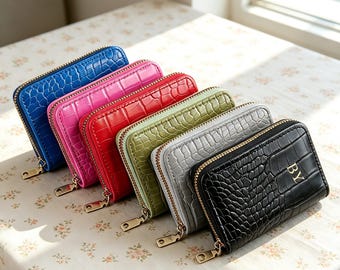Puede incluir: Una fila de seis carteras rectangulares con cremallera y textura de cocodrilo, dispuestas en línea diagonal. Las carteras son azul, rosa, roja, verde, gris y negra. La cartera negra presenta las letras doradas BY en la parte frontal.