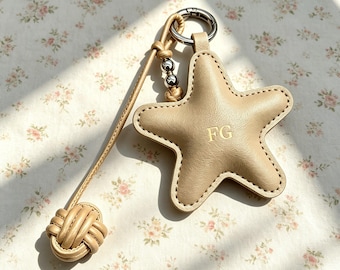 Puede incluir: Un llavero de cuero beige con forma de estrella que presenta las letras FG grabadas en el centro. El accesorio incluye una anilla de metal plateado, dos pequeñas cuentas plateadas y un largo cordón de cuero con un nudo decorativo.