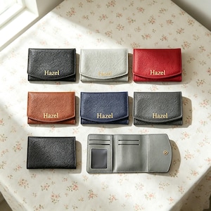May include: A collection of eight small, textured card holders in black, light gray, red, brown, navy blue, and dark gray. Each closed holder features the name Hazel embossed in gold. One light gray holder is shown open, revealing card slots.