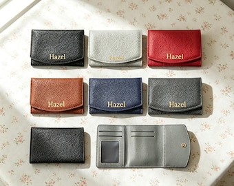 Puede incluir: Una colección de ocho tarjeteros pequeños y texturizados en negro, gris claro, rojo, marrón, azul marino y gris oscuro. Cada estuche cerrado lleva el nombre Hazel grabado en oro. Un estuche gris claro se muestra abierto, revelando ranuras.