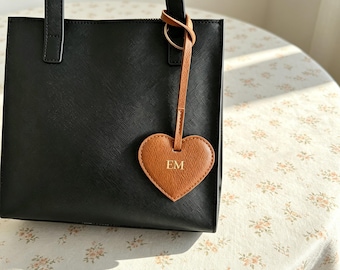 Puede incluir: Un bolso negro estructurado con dos asas verticales y una superficie texturizada. Una etiqueta de equipaje en forma de corazón de color canela cuelga de una asa, con las letras EM doradas en relieve en el centro del corazón.