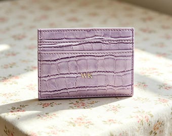 Puede incluir: Un tarjetero de color morado claro con un patrón de textura de piel de cocodrilo y las iniciales WK grabadas en dorado en la parte frontal. El accesorio rectangular cuenta con ranuras horizontales para tarjetas y costuras visibles en los bordes.