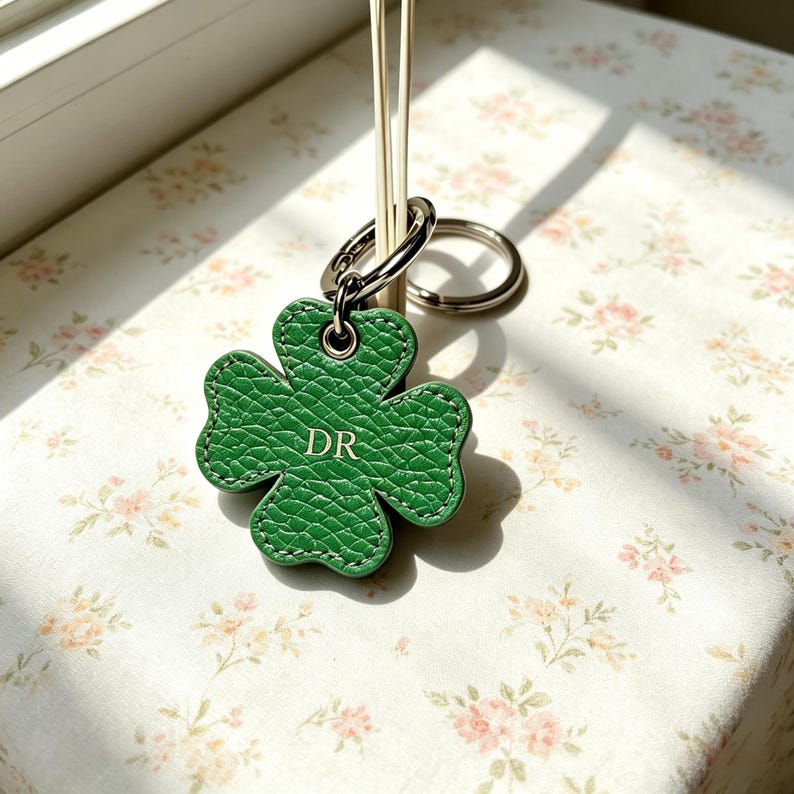 May include: A green four-leaf clover keychain made of textured material with white stitching along the edges. The center of the clover features the embossed letters DR in a gold-colored font. It is attached to a silver-toned metal ring.