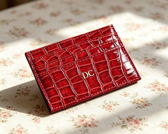 Puede incluir: Un tarjetero rectangular de color rojo intenso brillante con un patrón texturizado de piel de cocodrilo. La parte frontal presenta las letras DC en relieve de color dorado centradas en la superficie. El artículo tiene varias ranuras.