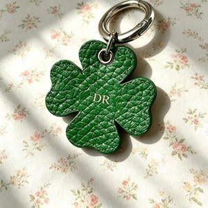 May include: A green four-leaf clover keyring made of textured leather with visible stitching along the edges. The centre of the clover features the gold-coloured letters DR embossed on the surface. A silver-toned metal ring is attached to the top.