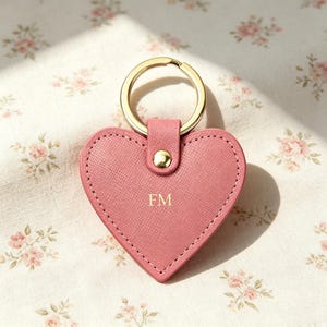 May include: A heart-shaped keychain made of pink textured material with gold-tone stitching around the edges. It features a gold-tone metal ring attached by a small tab with a gold-tone rivet. The letters FM are embossed in gold in the center.