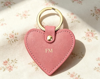 May include: A heart-shaped keychain made of pink textured material with gold-tone stitching around the edges. It features a gold-tone metal ring attached by a small tab with a gold-tone rivet. The letters FM are embossed in gold in the center.