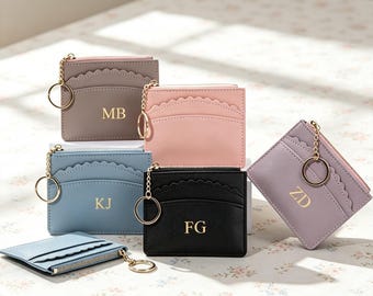 Puede incluir: Una colección de tarjeteros rectangulares en gris, rosa, azul, negro y lavanda, cada uno con un borde superior festoneado, cremallera dorada, llavero dorado e iniciales grabadas en oro como MB, KJ, FG y ZD.