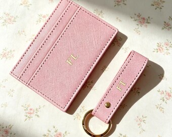 Puede incluir: Un tarjetero rectangular de color rosa claro y un llavero rectangular a juego con una anilla de metal dorado. Ambos artículos presentan una superficie texturizada y están personalizados con las letras doradas FL en el tarjetero y FT en el llavero.