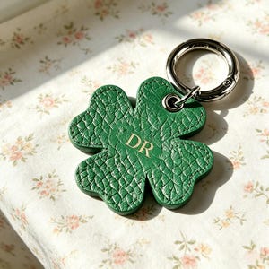 May include: A clover-shaped keychain crafted from textured green leather, featuring the gold-embossed letters DR in the center. The charm is attached to a polished silver-tone metal split ring, providing a secure way to organize keys or accessories.