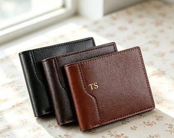 Puede incluir: Tres carteras plegables en negro, marrón oscuro y marrón rojizo, cada una con un acabado texturizado y bordes cosidos. La cartera marrón rojiza del frente muestra las iniciales TS grabadas en color dorado en el panel frontal derecho.