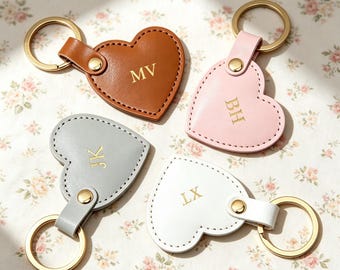 May include: Four heart-shaped keychains made of smooth material in brown, light pink, gray, and white. Each keychain features a gold-toned metal ring, a matching gold-toned snap button, and gold-embossed initials: MV, BH, JK, and LX respectively.