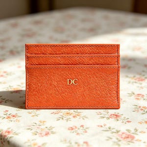 May include: A rectangular orange leather card holder with a textured grain finish and visible stitching along the edges. The front features two horizontal card slots and the gold embossed capital letters DC centered on the main pocket surface.