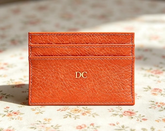 Puede incluir: Un tarjetero rectangular de cuero naranja con un acabado de grano texturizado y costuras visibles en los bordes. La parte frontal presenta dos ranuras horizontales para tarjetas y las letras mayúsculas doradas DC centradas.