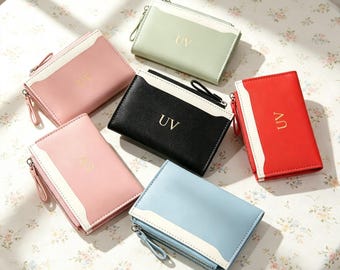 Puede incluir: Un conjunto de seis carteras rectangulares en rosa pastel, verde salvia, negro, rojo brillante y azul claro. Cada cartera presenta un panel blanco, un cierre de cremallera lateral con tirador a juego y las letras UV grabadas en oro.