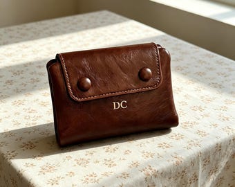 Puede incluir: Una cartera de cuero marrón oscuro con una solapa plegable, dos botones circulares a juego y las letras DC grabadas en color dorado en el centro de la parte frontal. La cartera tiene un acabado liso ligeramente texturizado y forma rectangular.