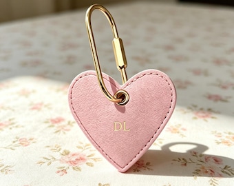 Puede incluir: Un llavero en forma de corazón de color rosa claro con una superficie texturizada y herrajes de metal dorado. El llavero está sujeto a un mosquetón dorado y tiene grabadas las letras mayúsculas DL en el centro del corazón.