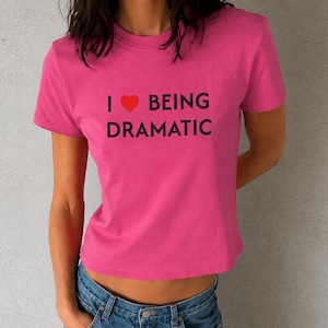 Puede incluir: Una camiseta de manga corta de color rosa brillante con el texto negro "I" seguido de un símbolo de corazón rojo y las palabras "BEING DRAMATIC" impresas en una fuente negrita sin remates en el centro del pecho.