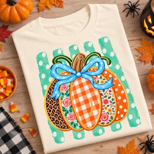 Può includere: Una t-shirt color crema con una grafica centrale raffigurante una zucca decorata con vari motivi, tra cui stampa leopardata, quadretti vichy arancioni e disegni floreali. Un fiocco blu brillante sovrasta la zucca su uno sfondo a pois color ottanio.