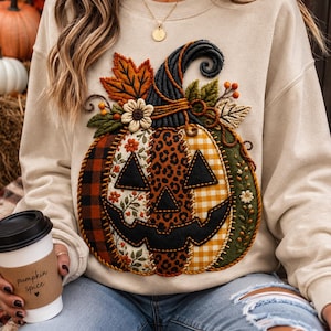 Peut inclure: Un sweat-shirt beige à col rond orné d'une grande applique de citrouille texturée et à motifs multiples sur le devant. Le motif de la citrouille comprend des sections à carreaux noirs et rouges, à fleurs, à imprimé léopard et à vichy jaune.