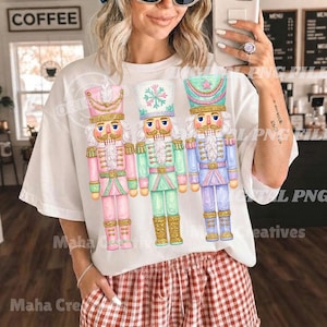 Op de afbeelding: Een wit t-shirt met een grafische print van drie notenkrakerfiguren in pastelkleuren. De linker notenkraker is roze, de middelste is mintgroen met een sneeuwvlokontwerp op de hoed en de rechter is lichtblauw, allemaal met gouden accenten.